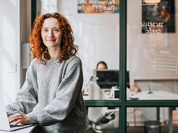 Rödhårig kvinna på kontor ser leendes in i kameran